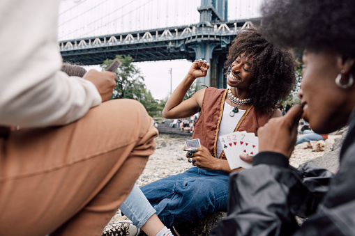 Multi-ethnic group of friends resting and enjoying a break next to Brooklyn Bridge in New York. They are playing with cards sitting on the rocks by the river.