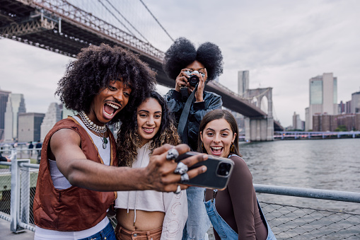 A multi-ethnic group of friends taking a selfie in Brooklyn with Brooklyn Bridge and lower Manhattan on the background. This is a famous tourist spot for a visit in New York City.