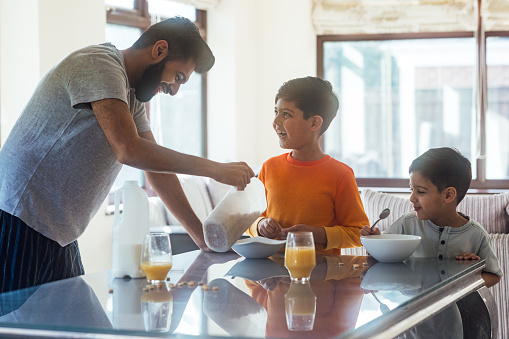 A medium close up of a father and his young sons as they prepare and eat their breakfast cereal. They are up early before their mother in their family home in Newcastle Upon Tyne in the North East of England.He is pouring the milk in the cereal for them.