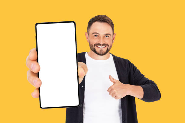 istock Handsome young man holding a smartphone mockup with a blank screen and smiling on an orange background. 1744759298