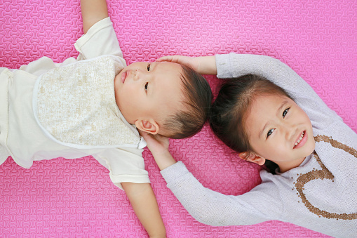 Adorable cute Asian sister and little brother lying on pink mattress mat looking at camera.