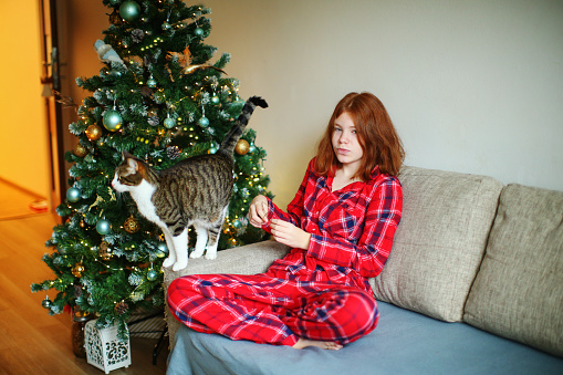 girl with red pajamas holds a cat. Christmas tree in the background.