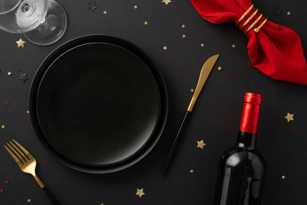 istock Premium restaurant New Year's special. Top view of table setup including plates, tableware, red napkin and holder, wine bottle, glass, glistening confetti against black background with space for text 1744839716