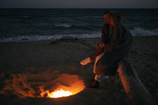 Couple sitting together with tenderness near campfire on the beach at the evening focus on the sea