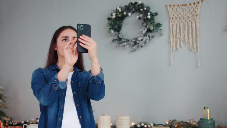 Concerned upset young woman in a Christmas-decorated kitchen puzzled over malfunctioning phone, facing loss of WiFi, 3G cellular coverage and data during the festive New Year and Christmas season.