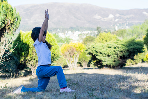 Little Latin girl practices yoga in the park, with a background full of mountains and beautiful bright green bushes.