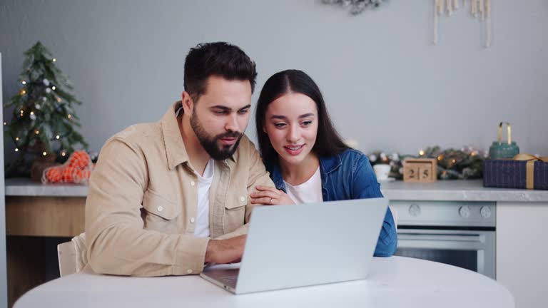 Cheerful Spanish Pair Engaging in Online Shopping Amidst a Lushly Decorated Christmas Kitchen Multiracial Couple Enthusiastically Choosing Ordering Festive Gifts on Laptop, Emboding Holiday Spirit.