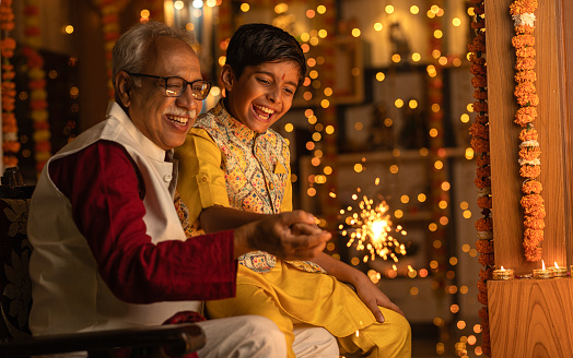 Grandfather holding cheerful grandson's hand and playing with sparkler while sitting at home during Diwali festival