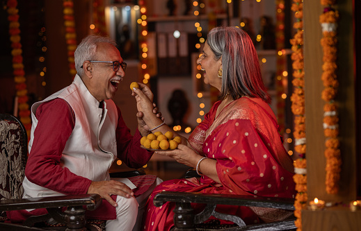 Senior wife dressed in sari feeding laddoo to cheerful husband while sitting on chairs and celebrating Diwali festival together at home