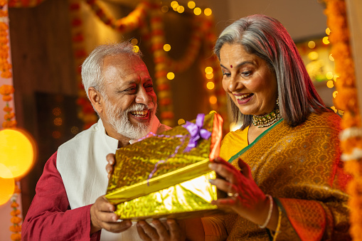 Happy senior husband giving surprise gift to excited wife in illuminated home on occasion of Diwali festival