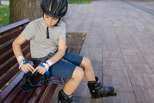 Young male skater sitting in the park and preparing for roller skating. He is tying his inline skates and putting on his protective skating equipment.