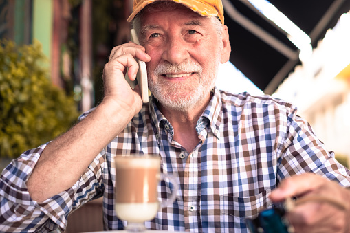 Senior man with cap holding talking on mobile phone sitting outdoor at a cafe table having break with cappuccino drink, relaxed retirement lifestyle