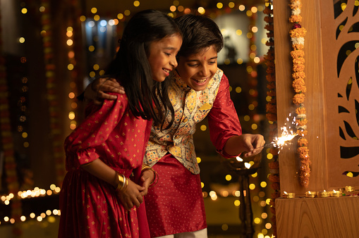Smiling boy with hand on sister's shoulder burning sparkler while standing in illuminated home during Diwali festival