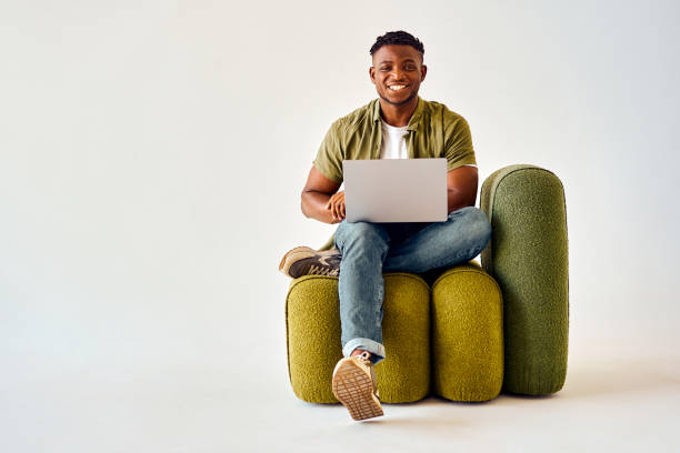 chico afroamericano feliz sentado en una silla de diseño y sosteniendo una computadora portátil inalámbrica. - red chairs fotografías e imágenes de stock