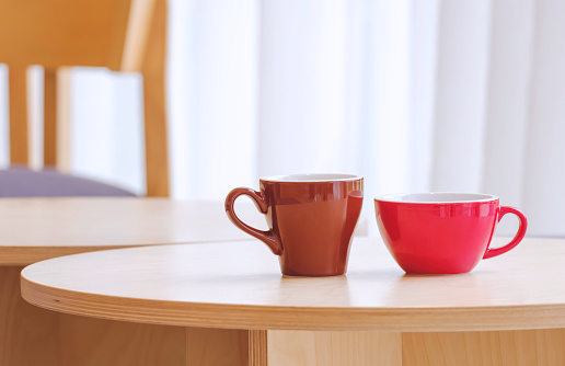 Two red and brown coffee mug on round wooden table with blurred background of white curtain in modern living room, close up with copy space