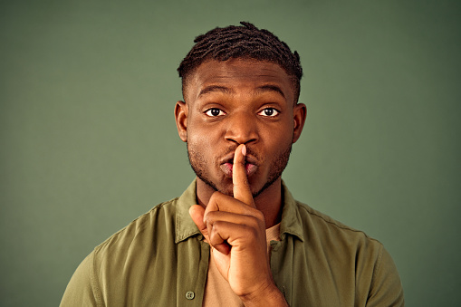 Portrait of serious young man with afro hairstyle doing shush gesture near lips while standing over green background. Casually dressed black guy tying to keep conspiracy in studio.