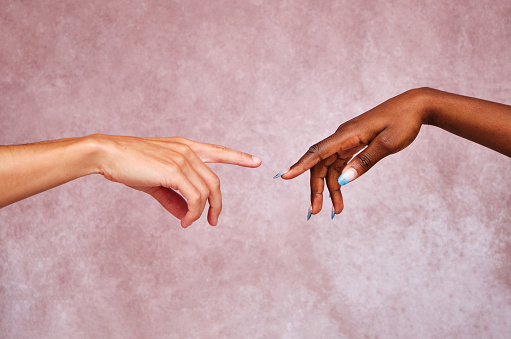 Hand of a Caucasian man reaching out with an extended finger towards the hand of an Afro American woman with blue fingernails on a pink background.