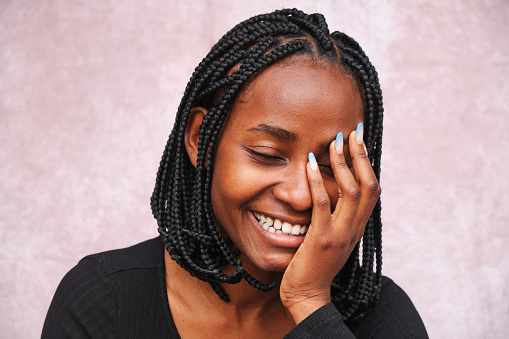 Portrait on a brown background of Afro woman laughing, eyes closed and a hand on her face with long nails painted blue.