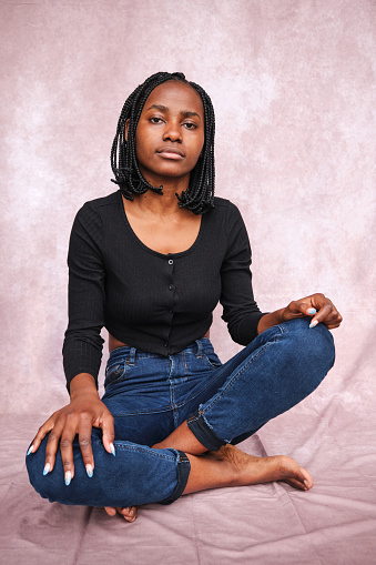 Afro woman sitting on the studio floor on a brown background looking at the camera with her legs crossed and hands resting on her knees.