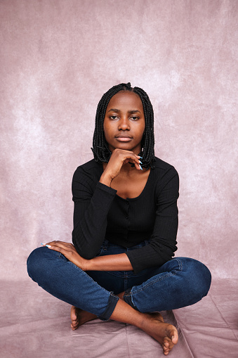 Afro-American woman looking into the camera, sitting on a brown background with her legs crossed and her feet bare, resting her chin on her hand.