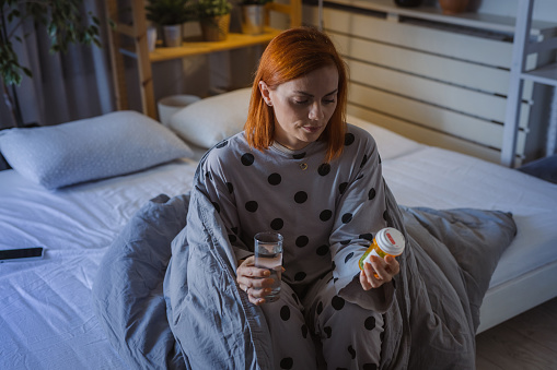 Sad mid adult woman sitting on the bed during the night. She is holding glass of water and bottle of medicines
