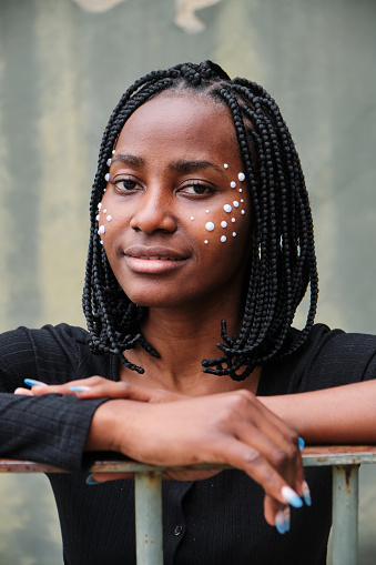 Portrait of an African woman with braids in her hair and beads on her face, looking into the camera and leaning against a metal fence.