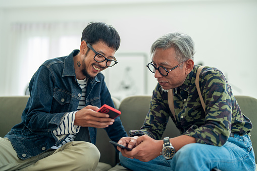 Asian Man and Senior Father Enjoying Quality Family Time Using Smartphones in Cozy Living Room.