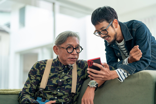 Multi-Generation Connection: Asian Freelancer and Elderly Father Share Digital Moments at Home.
