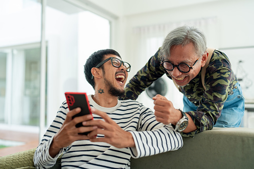 Intergenerational Bonding: Casual Moments of Happiness as Asian Man and Senior Father Connect Through Technology in Their Living Room.