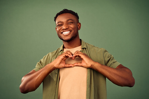 African american handsome guy smiling sincerely and making heart shape with hands over green background. Young male person in khaki shirt showing romantic feelings in studio.
