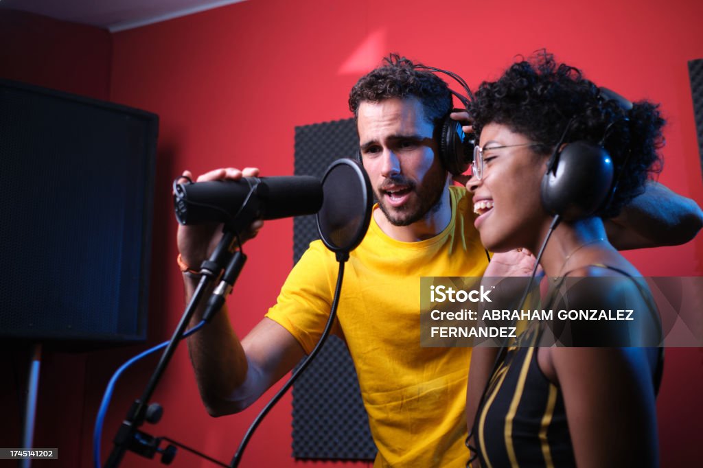 Caucasian man singing in a recording studio next to a curly haired African woman. Caucasian man holding microphone with one hand and wearing headphones is singing in a recording studio next to a curly haired African woman wearing. glasses and headphones. The Media Stock Photo