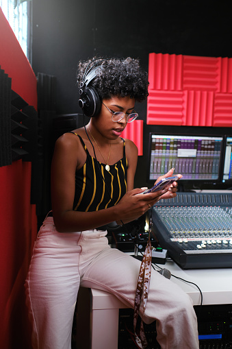Afro woman leaning on the recording studio table, consulting her cell phone while listening to the recording through headphones.