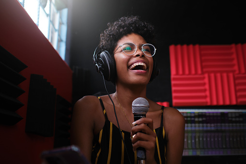 Portrait of a hispanic woman wearing headphones and holding a microphone with one hand while happily singing in a recording studio.
