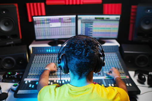 Rear view of the sound engineer sitting in front of the mixing desk with headphones on and modifying the sound.