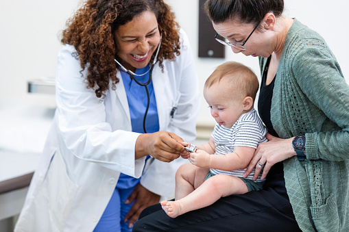 The mid adult female doctor laughs as the little baby held by her mother plays with the stethoscope during the medical examination.