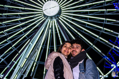 Young happy couple smiling looking at camera with the lights of a ferris wheel in the background