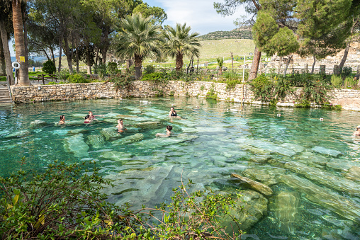 Pamukkale, Denizli, Turkey - March 31, 2023. The Antique Pool at Hierapolis ancient site in Pamukkale, Turkey. The sacred pool in the courtyard of this spa has submerged sections of original fluted marble columns. The water is abundant in minerals. View with people in and around the pool.