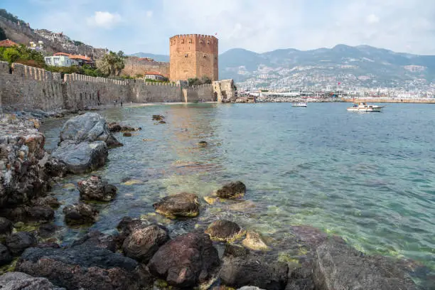 View toward old harbour walls and Red Tower in Alanya, Turkey. The five-storey octagonal defence tower was constructed in 1226 by Seljuk Sultan Alaeddin Keykubad I, and currently houses the Ethnographic Museum. Measuring nearly 30m in diameter, more than 30m in height, it has a central cistern within for water storage, and looms over the harbour at the lower end of Iskele Caddesi. View with unidentifiable figures of people.