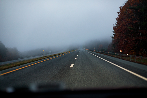 View through the windshield of a car speeding in complete solitude along an early morning foggy, hazy, hilly New Hampshire expressway in October.