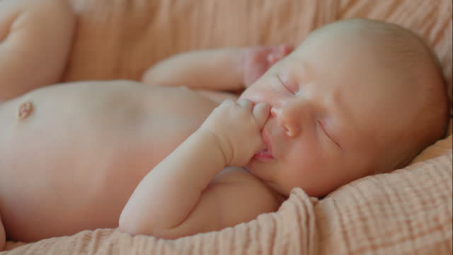 A newborn baby is captured in a close-up shot, lying peacefully in a cradle and gently sucking on his tiny finger. After couple of seconds he falls asleep. This heartwarming footage encapsulates the precious early moments of a new life, radiating a feeli