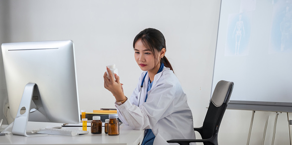 Female doctor writing in a notebook and holding a medicine bottle works at a computer while there were many medicine bottles on the table while sitting at a work desk.