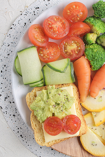 Light lunch or dinner with cherry tomato, cucumber, zucchini, carrot, broccoli, radish and toasted bread with avocado and cheese served on a white table.