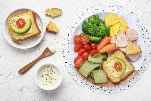 Light lunch or dinner with cherry tomato, cucumber, zucchini, carrot, broccoli, radish and toasted bread with avocado and cheese served on a white table.