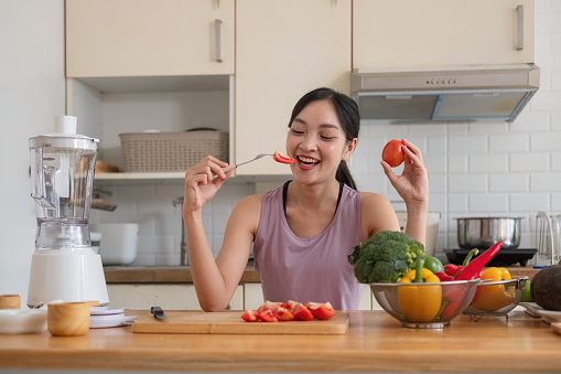 Young Asian healthy beautiful woman with casual clothes is smile and slicing cut tomatoes to eat fruit to diet at home in kitchen .