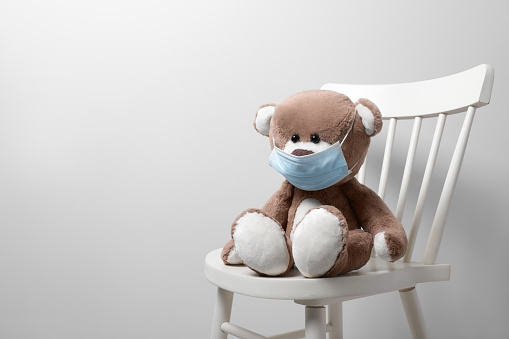 Cute teddy bear in medical mask on chair near light wall, space for text