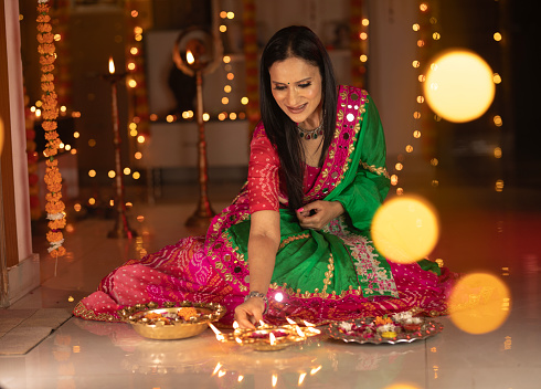 Smiling beautiful woman dressed in traditional clothing sitting with diyas and plates on floor in illuminated home during Diwali festival