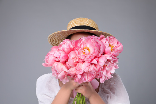 Young woman covering her face with bouquet of peonies on grey background