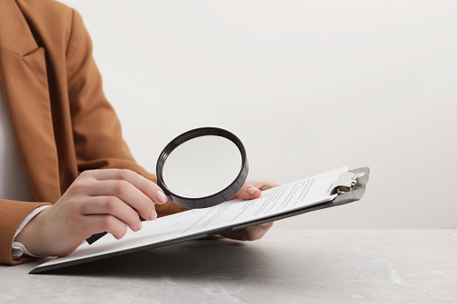 Woman looking at document through magnifier at light gray table, closeup. Searching concept