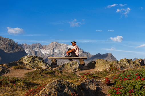 Hiker rests on a wooden bench on top of a mountain ridge overlooking the alpine peaks of Bernese Alps, Aletsch glacier, UNESCO World Heritage Site, Valais Canton, Switzerland