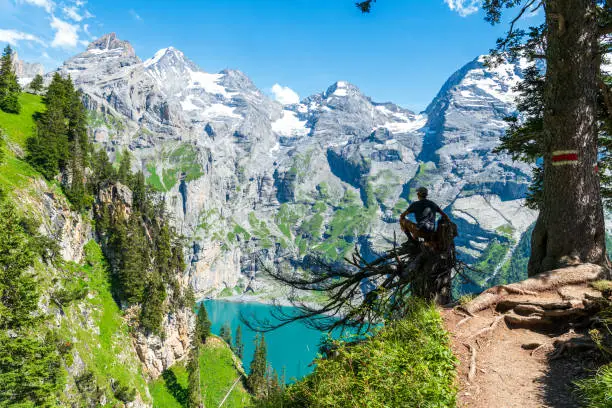 View of a male hiker enjoying the view of the turquoise lake of Oeschinensee surrounded by rocky peaks with glaciers, Kandersteg, Bern Canton, Switzerland